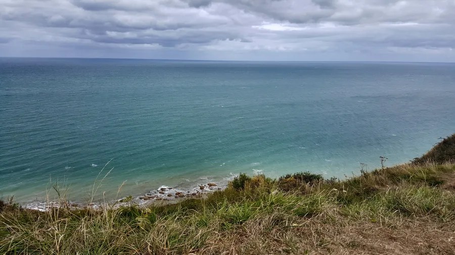 Panoramaansicht des Meeres, mit sanften Wellen und einem bewölkten Himmel über der Küste, umgeben von grünem Gras.