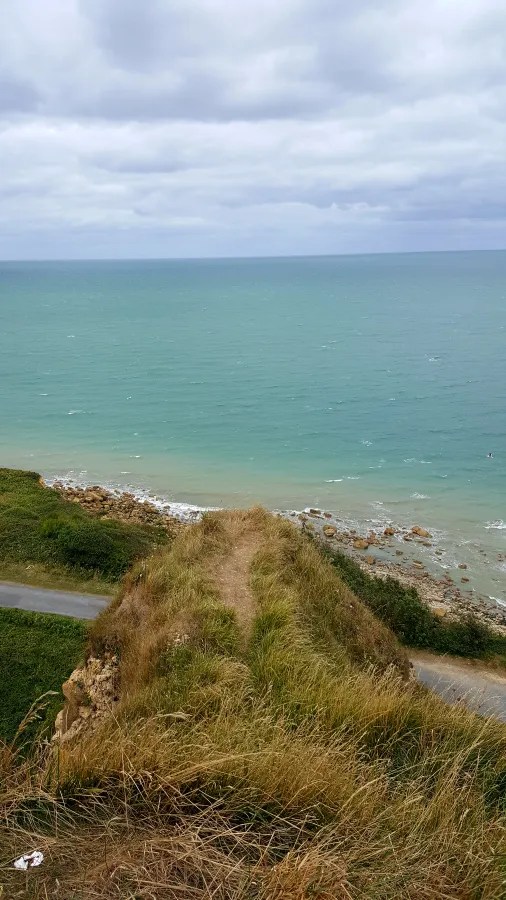 Blick vom Kliff auf die ruhige Küste mit sanftem, grün-blauem Wasser und bewachsenen Hängen in der Normandie.