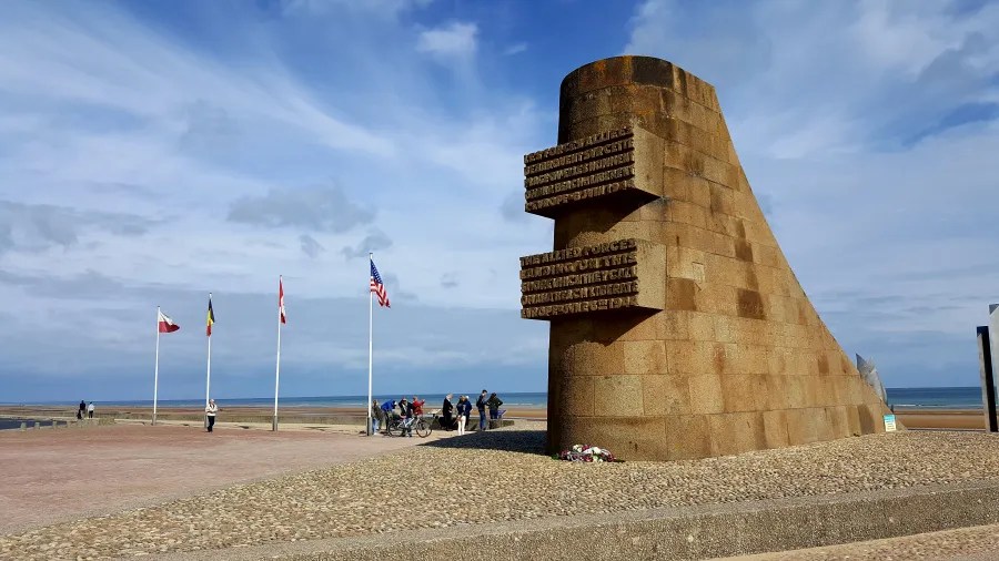 Denkmal am Strand mit mehreren Nationalflaggen im Hintergrund und einer Gruppe von Besuchern.