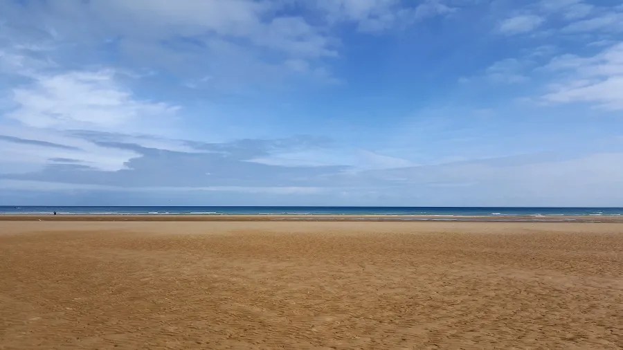 Weitläufiger Sandstrand mit sanften Wellen und leicht bewölktem Himmel über dem Meer.