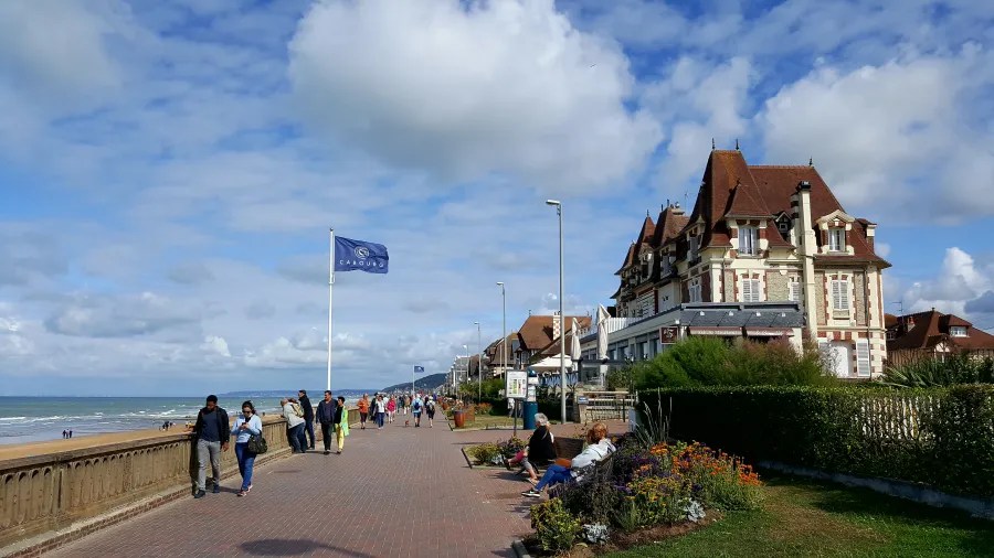Blick auf die Promenade von Cabourg, Frankreich, mit Besuchern, die entlang des Meerufers spazieren, und typischer Architektur im Hintergrund unter einem bewölkten Himmel.