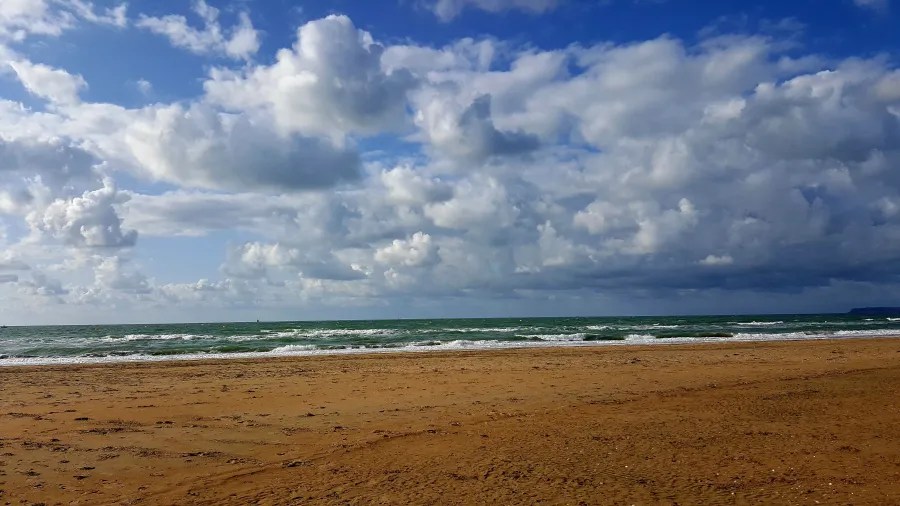 Ein weitläufiger Sandstrand mit sanften Wellen und einem dramatischen Himmel voller Wolken, der eine ruhige Küstenlandschaft darstellt.