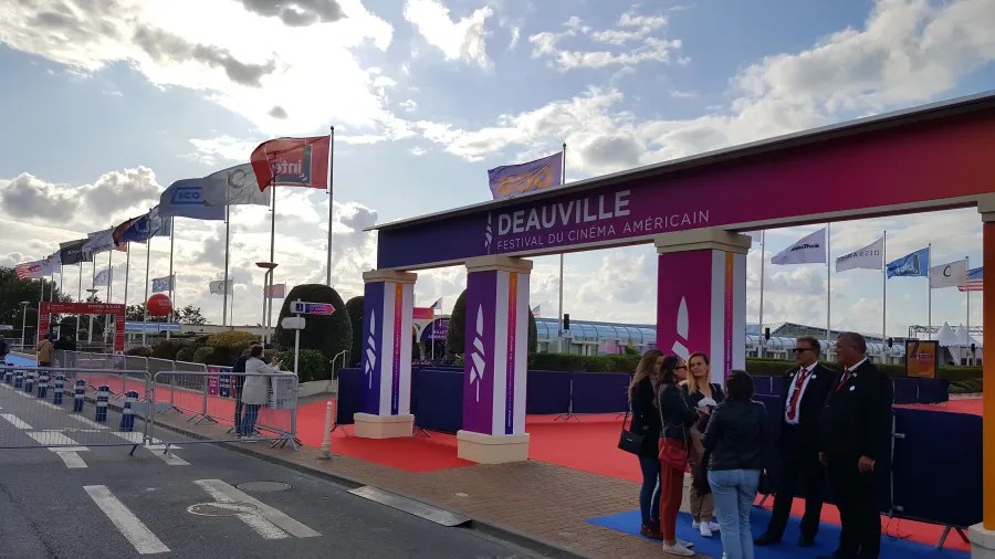 Eingang zum Festival du Cinéma Américain in Deauville, mit roten Teppich, Sicherheitskräften und mehreren Fahnen vor einer Wolkenlandschaft.