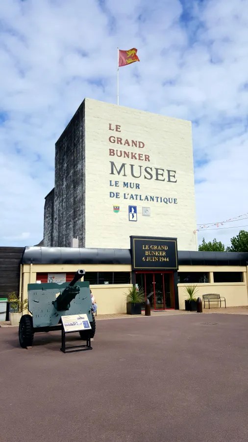 Das Bild zeigt das Grand Bunker Museum in der Normandie, Frankreich, mit einer großen weißen Wand und einem Kanonenmodell im Vordergrund. Eine Flagge weht über dem Gebäude, das die Geschichte des Atlantikwalls thematisiert.