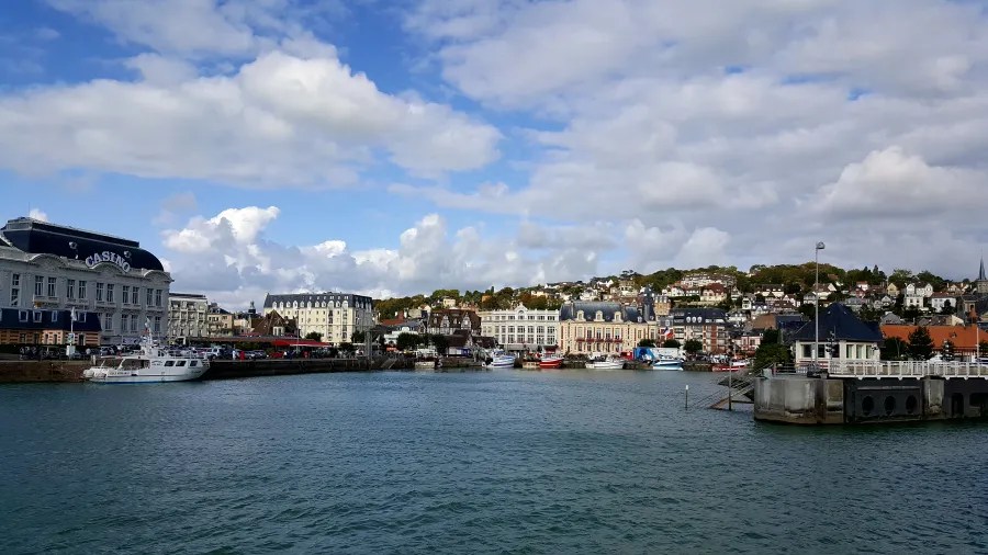 Blick auf den Hafen von Cabourg mit zahlreichen Booten, einem Casino und malerischen Gebäuden im Hintergrund unter einem wolkigen Himmel.