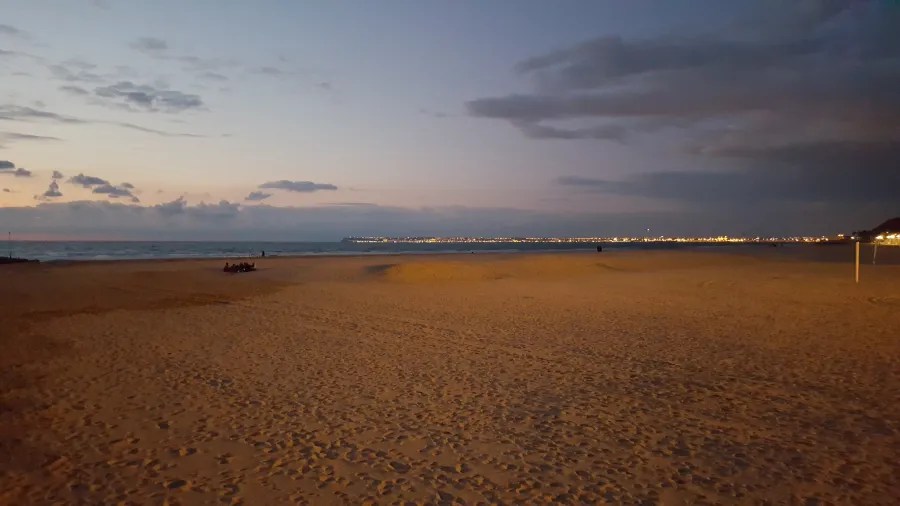 Bei Dämmerung zeigt sich der leere Strand mit sanftem, goldenem Sand und den Wellen des Atlantiks, während die Stadtlichter im Hintergrund langsam erleuchten.