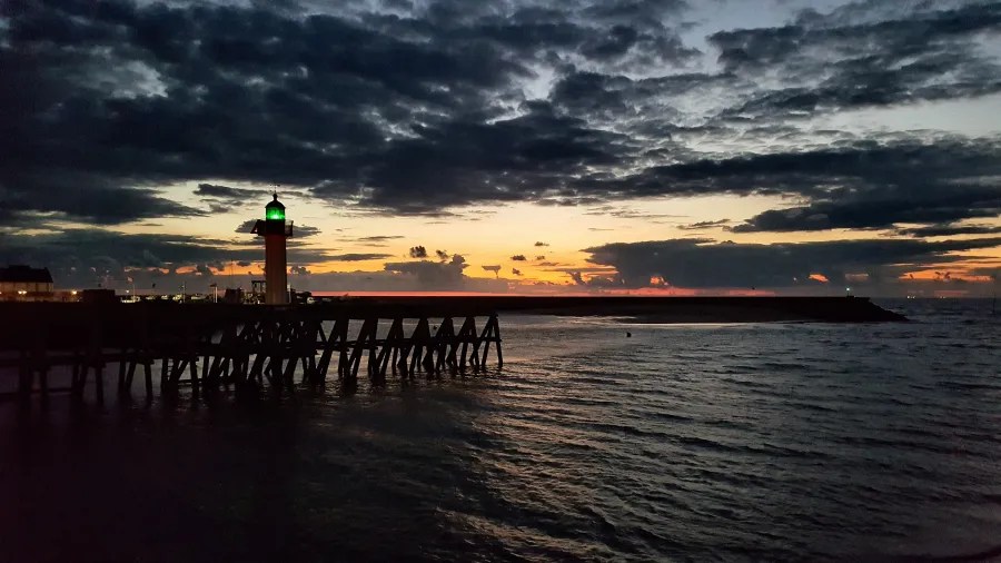 Sonnenuntergang über dem Meer mit einem Leuchtturm und wolkenverhangenen Himmel.