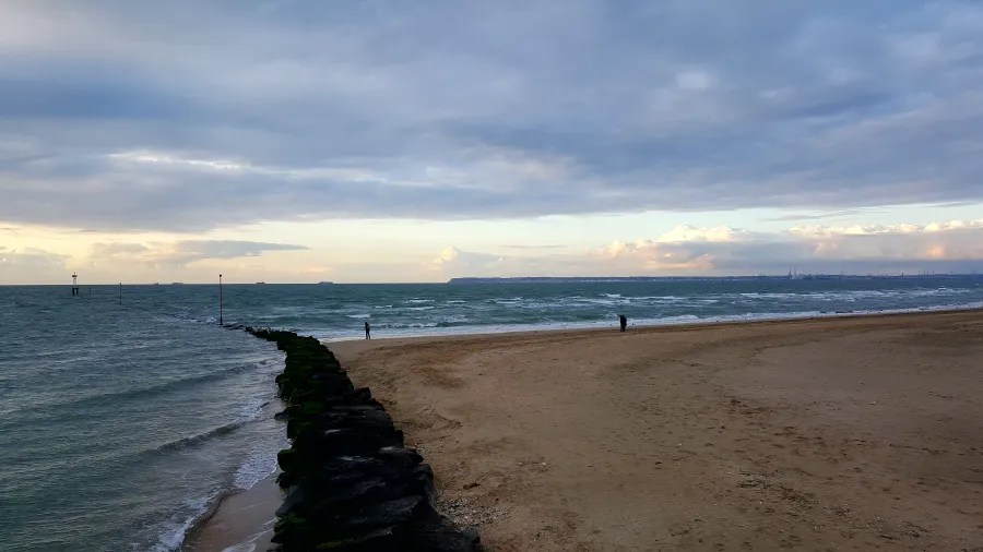 Blick auf den Strand und das Meer mit einer Person, die am Wasserrand steht, während sich Wolken über dem Himmel ziehen.