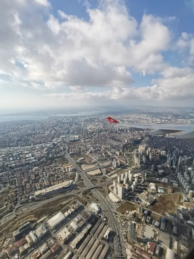 Luftaufnahme einer Stadt mit vielen Gebäuden und Straßen, gesehen aus einem Flugzeugfenster, im Hintergrund blaue Himmel mit Wolken und das Flugzeugflügel mit einem roten Logo.