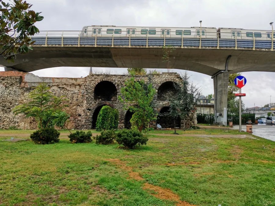 Eine alte, teilweise bewachsene Mauer mit Bögen, im Vordergrund ein grüner Rasen mit kleinen Büschen, darüber eine moderne metroähnliche Brücke und ein Verkehrsschild.