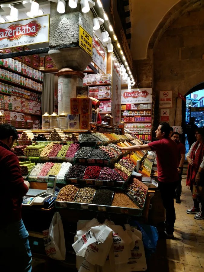 Ein Basar in Istanbul mit einer bunten Auswahl an Süßigkeiten und Gewürzen, beleuchtet von warmem Licht. Im Vordergrund verkauft ein Verkäufer verschiedene Produkte, während Kunden im Hintergrund durch den Markt schlendern.