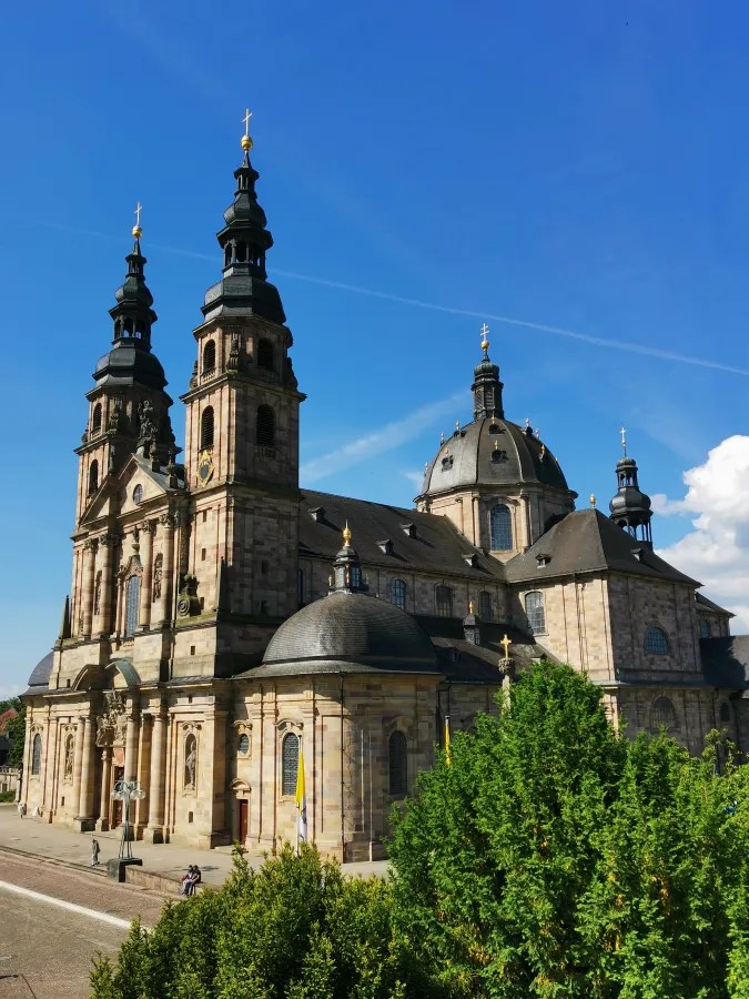 Blick auf den Fuldaer Dom, eine barocke Kirche mit markanten Türmen und einer Kuppel, umgeben von grünem Laub und klarem blauen Himmel.