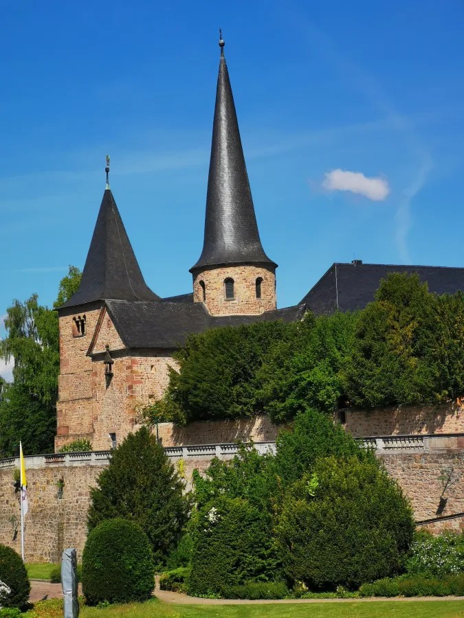 Blick auf die Michaelskirche in Fulda mit ihrem markanten, spitzen Kirchturm, umgeben von üppigem Grün und Bäumen an einem blauen Himmel.