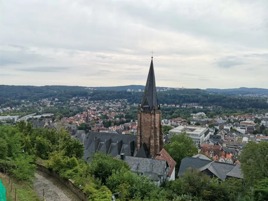 Panoramaaufnahme über Marburg mit Blick auf die Stadtkirche und die umliegende Landschaft. Der Kirchturm ragt in den Himmel und die Stadt ist umgeben von grünen Hügeln unter einem leicht bewölkten Himmel.