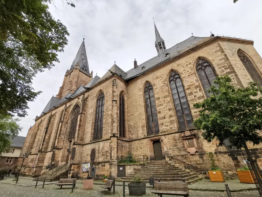 Die Ansicht der Elisabethkirche in Marburg, mit gotischen Türmen, umgeben von Bäumen und Stufen, unter einem bewölkten Himmel.