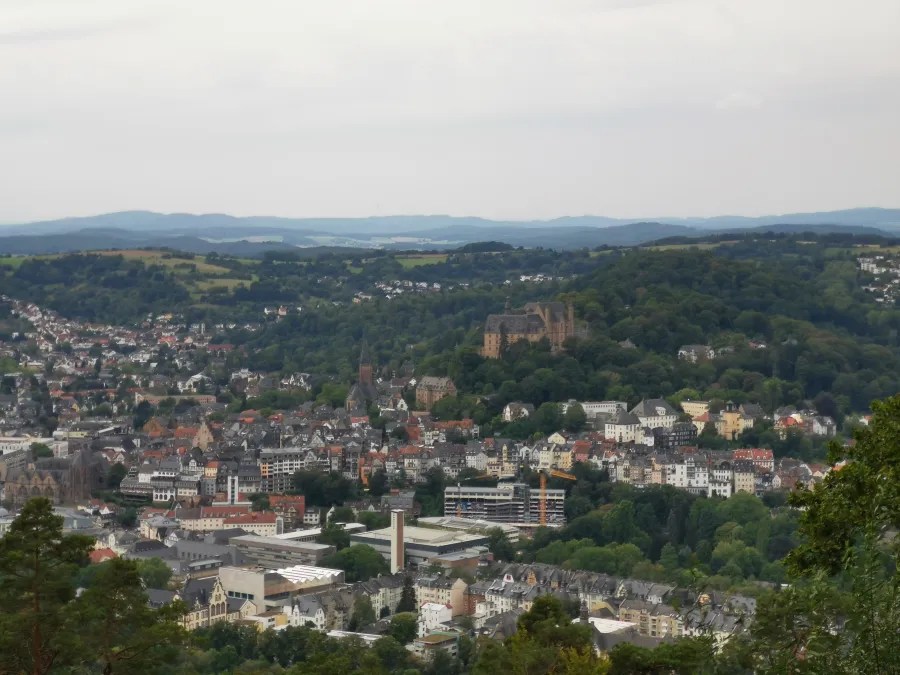 Blick auf Marburg mit der Landgrafenburg, umgeben von grünen Hügeln und historischen Gebäuden unter einem bewölkten Himmel.