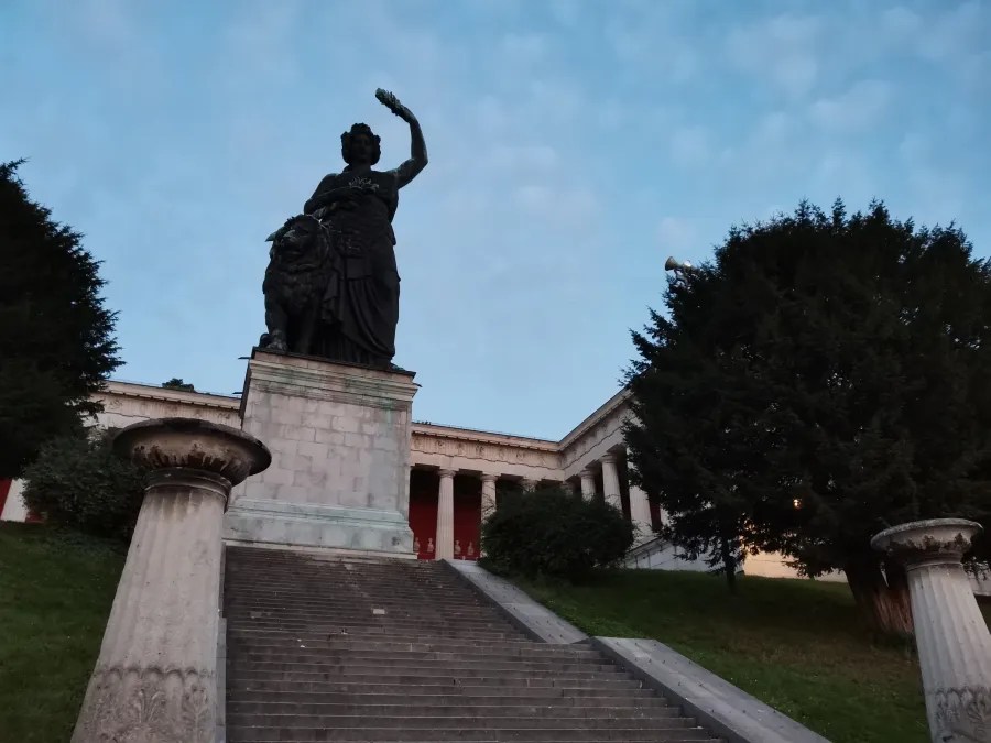Statue der Bavaria, die majestätisch auf einer Treppe vor einem Gebäude steht, umgeben von Bäumen und einem wolkigen Himmel.