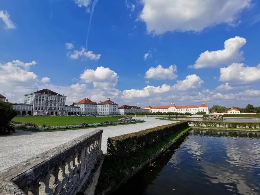 Blick auf das Schloss Nymphenburg in München mit blauen Himmel und weißen Wolken, umgeben von gepflegten Gärten und einem kleinen Wasserlauf im Vordergrund.