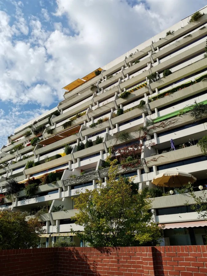 Modernes Wohngebäude mit zahlreichen Balkonen, bepflanzt mit Grünpflanzen und Sonnenschirmen, unter einem wolkenverhangenen Himmel.