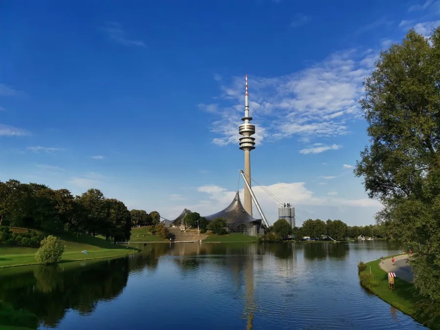 Panoramablick auf den Olympiaturm in München, umgeben von grünem Park und klarem Wasser unter blauem Himmel.
