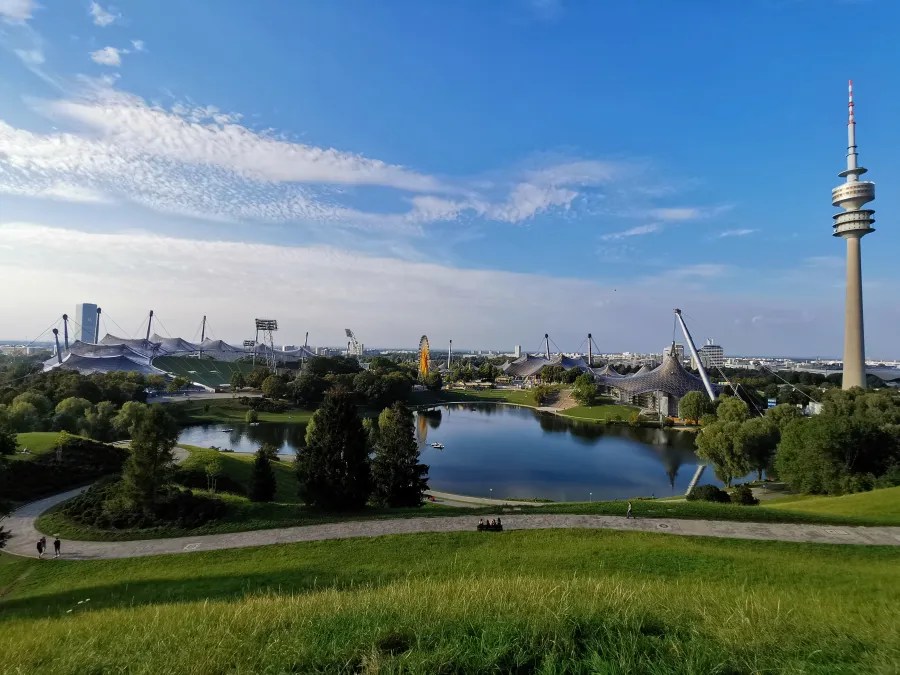Panoramablick auf den Olympiapark in München, mit einem ruhigen See im Vordergrund, umgeben von grünen Wiesen und Bäumen. Im Hintergrund sind die charakteristischen Zeltstrukturen des Olympiastadions und der Fernmeldeturm zu sehen, unter einem klaren blauen Himmel.