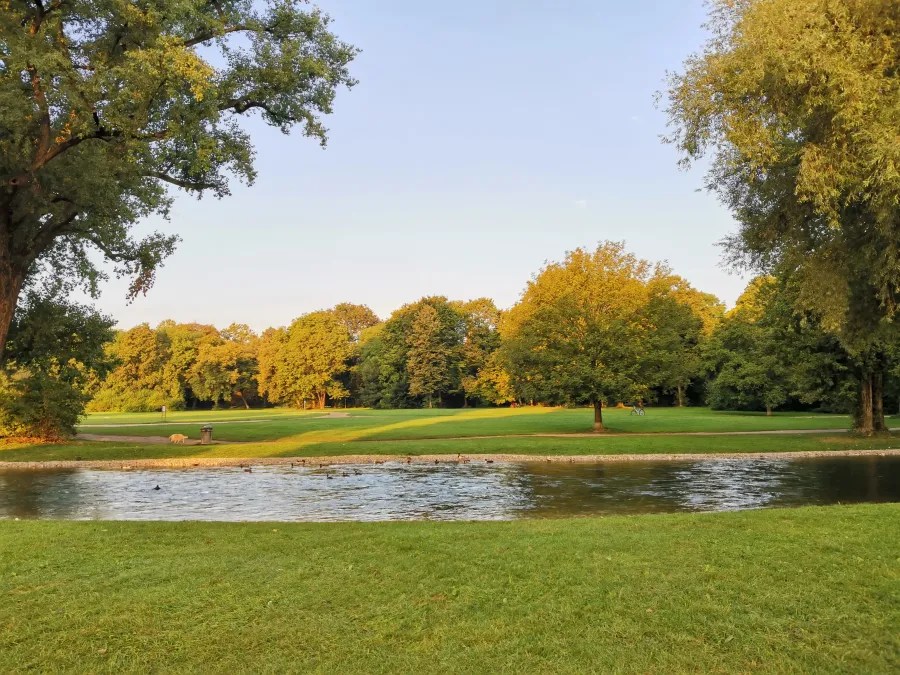 Ein ruhiger Park mit üppigem Grün, Bäumen und einem sanft fließenden Gewässer unter einem klaren Himmel.