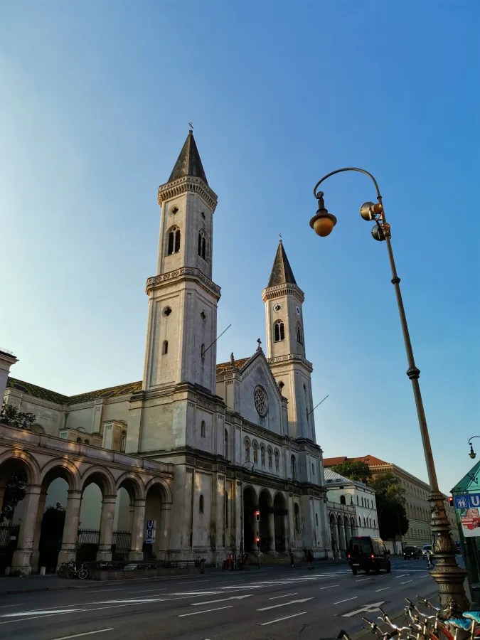 Blick auf eine historische Kirche mit zwei hohen Türmen und einer großen Fensterrose, umgeben von einer urbanen Landschaft und einem klaren blauen Himmel.