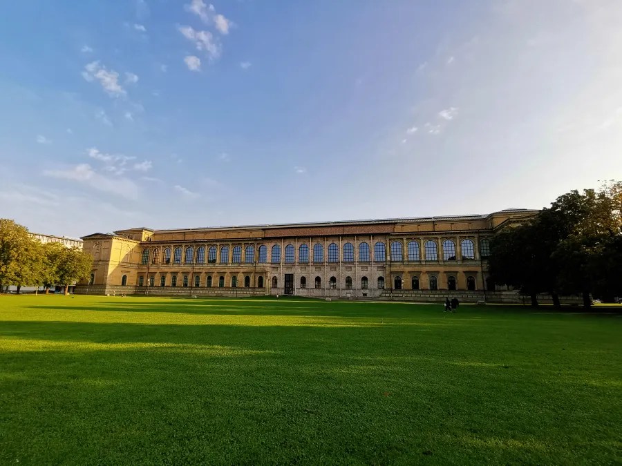 Das Gebäude der Alten Pinakothek in München mit einer weiten grünen Wiese im Vordergrund und einem klaren Himmel im Hintergrund.