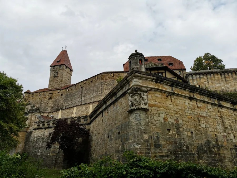 Blick auf die mächtige Veste Coburg mit ihrer dicken Steinmauer und Türmen, umgeben von Bäumen und einem bewölkten Himmel.