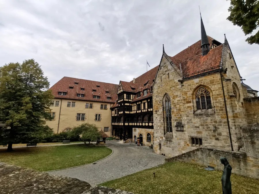 Eine historische Burg mit einem Mix aus Fachwerk und Steinarchitektur. Im Vordergrund ein Gartenbereich mit Bäumen und Bänken, im Hintergrund die Burg mit Spitzdächern und modernen Elementen.