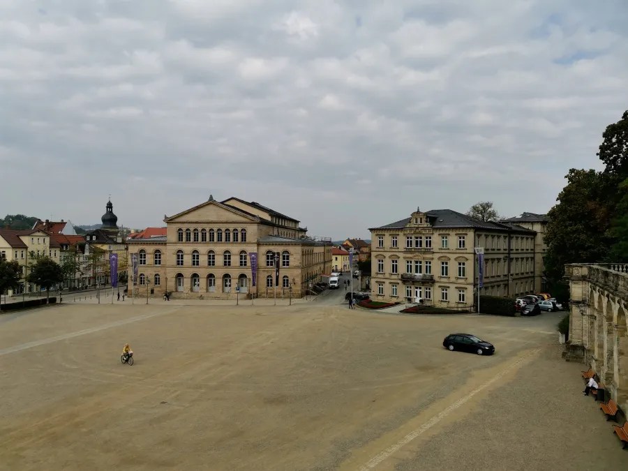Blick auf den Schlossplatz in Coburg mit dem Landestheater und angrenzenden Gebäuden, umgeben von einem Kiesplatz und leicht bewölktem Himmel.