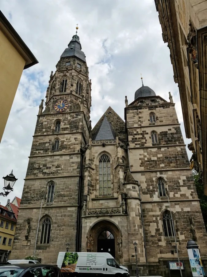 Die Fassade einer historischen Kirche in Coburg mit zwei Türmen, die eine Uhr zeigen. Ein grauer Lieferwagen steht vor dem Eingang, während der Himmel mit Wolken bedeckt ist.