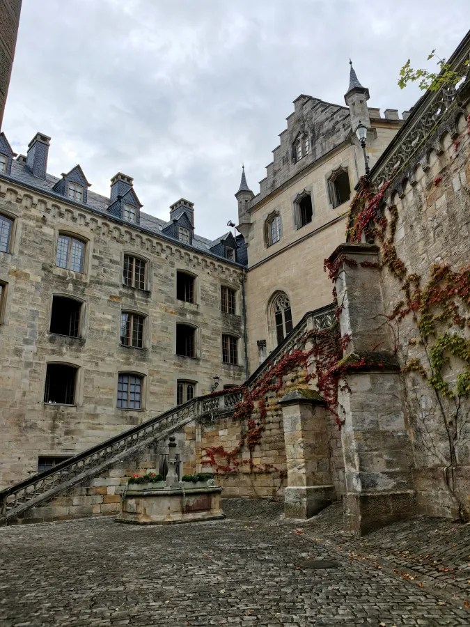 Innerer Bereich eines historischen Schlosses mit einer steinernen Treppe und einem Brunnen. Efeu rankt an der Wand. Die Architektur ist mittelalterlich und zeigt Fenster im gotischen Stil.