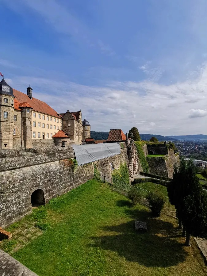 Blick auf die Veste Coburg mit historischen Gebäuden und grünen Wiesen im Vordergrund unter einem teilweise bewölkten Himmel.