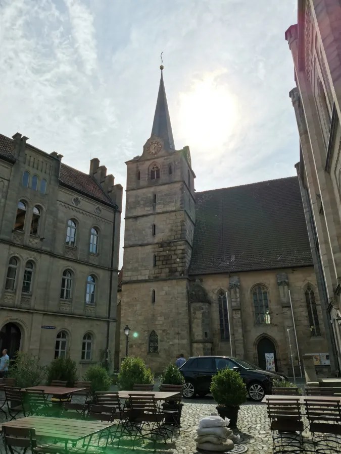 Blick auf den Turm einer Kirche in Coburg, umgeben von historischen Gebäuden und einem Platz mit Tischreihen.