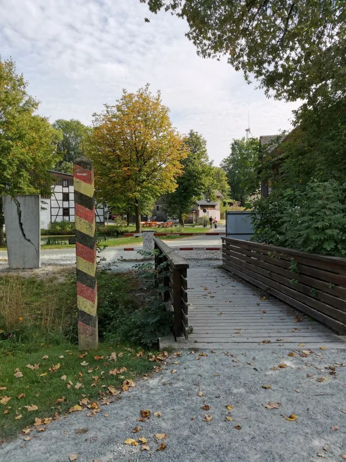Ein Bild des Tannbachs in Mödlareuth mit einem Holzsteg und grünen Bäumen, sichtbar sind auch Reste von Grenzmarkierungen mit bunten Streifen.
