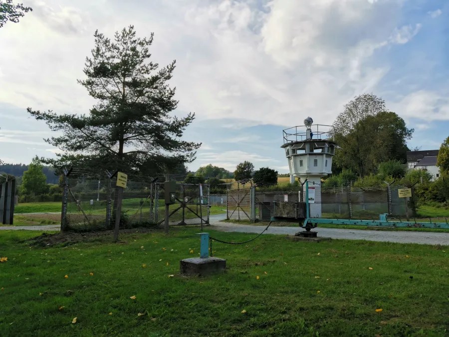 Blick auf einen ehemaligen Grenzposten mit Wachturm, umgeben von einem Zaun und Bäumen.