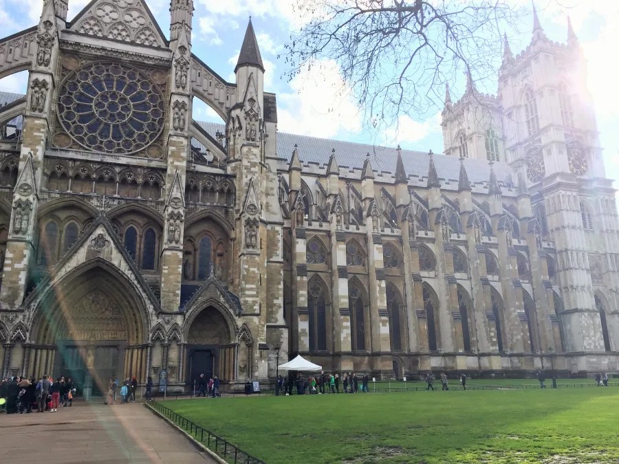 Die Fassade von Westminster Abbey, einem monumentalen Bauwerk in London, mit detaillierten gotischen Elementen und einer großen Rosette. Eine Menschenmenge versammelt sich im Vordergrund auf dem grünen Rasen.