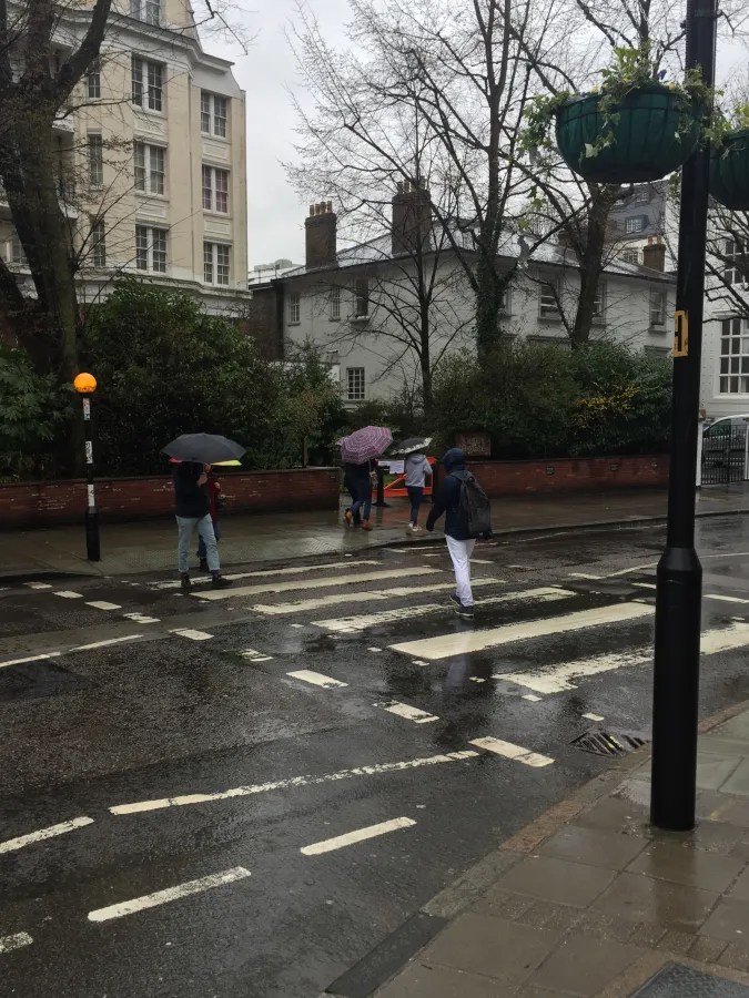 Personen überqueren den bekannten Zebrastreifen der Abbey Road bei Regen, während sie Regenschirme benutzen.
