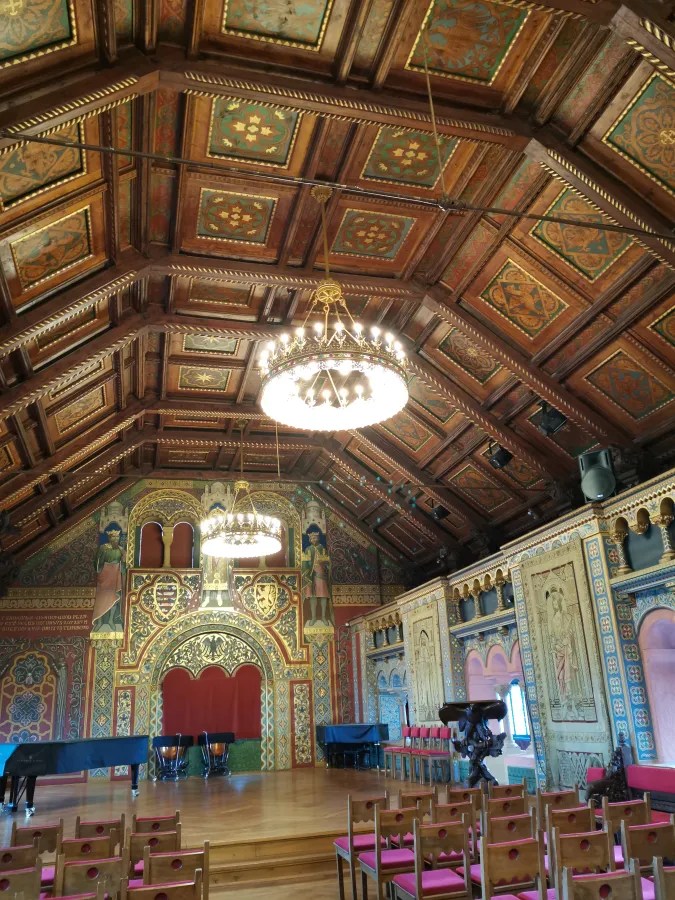 Interior view of an ornate hall with a high wooden ceiling, decorative chandeliers, and intricately painted walls featuring historical figures and designs. The space includes seating and a grand piano.