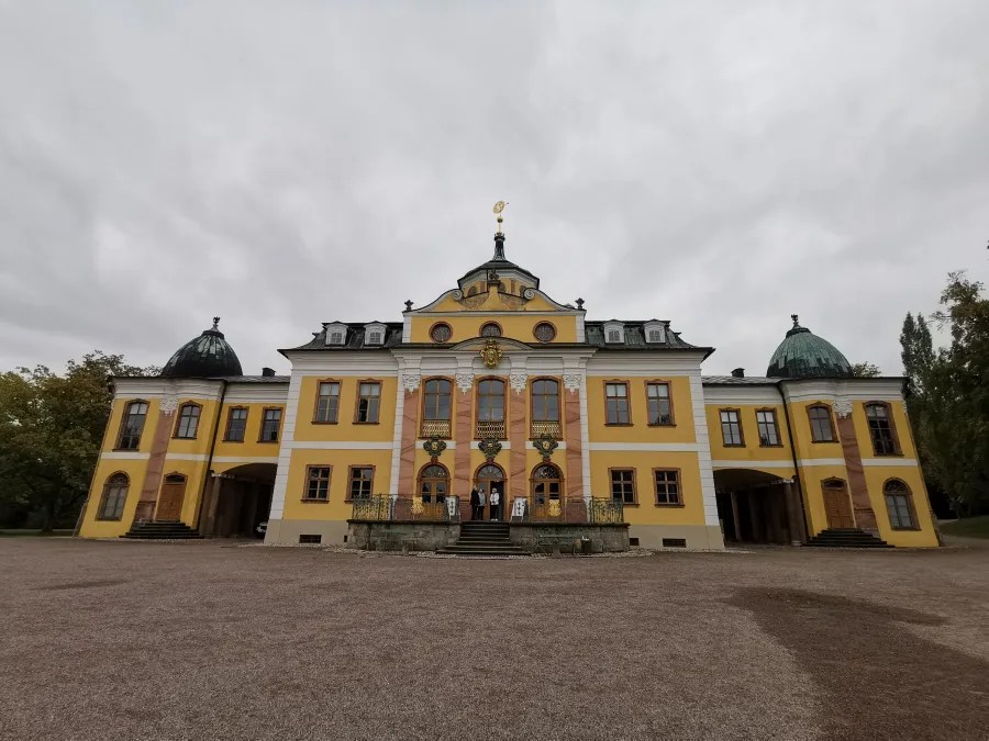 Das Schloss Belvedere in Weimar, eine gelb gestrichene historische Architektur mit grünen Kuppeln, umgeben von einem gepflasterten Vorplatz und Bäume im Hintergrund, unter einem bewölkten Himmel.