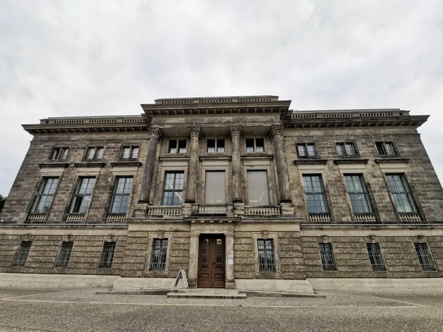 Die Vorderansicht eines historischen, monumental gestalteten Gebäudes mit Säulen und großen Fenstern, umgeben von einem grauen Himmel.