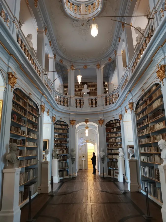 Innenansicht einer Bibliothek mit dekorativen Wänden, Regalen voller Bücher und Statuen von Autoren, die eine Person stehen sieht.