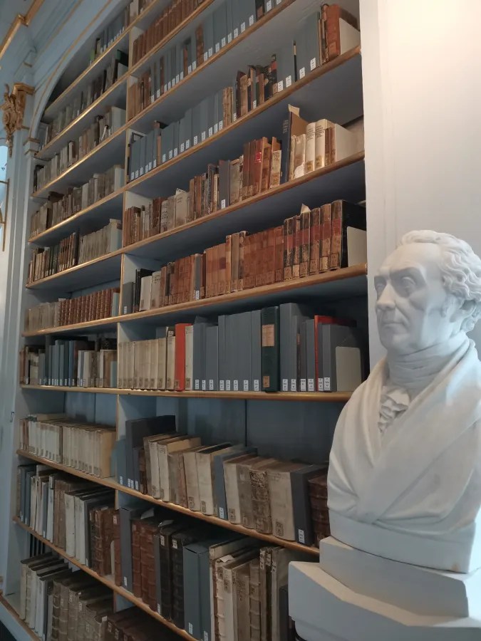 Blick auf ein Bücherregal in einer historischen Bibliothek, das mit alten und modernen Buchbänden gefüllt ist, neben dem eine weiße Büste eines Mannes steht.