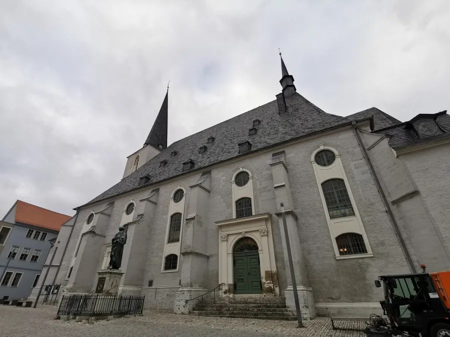 Außenansicht der Herderkirche in Weimar mit grauer Fassade, spitzem Dach und statue vor dem Eingang.