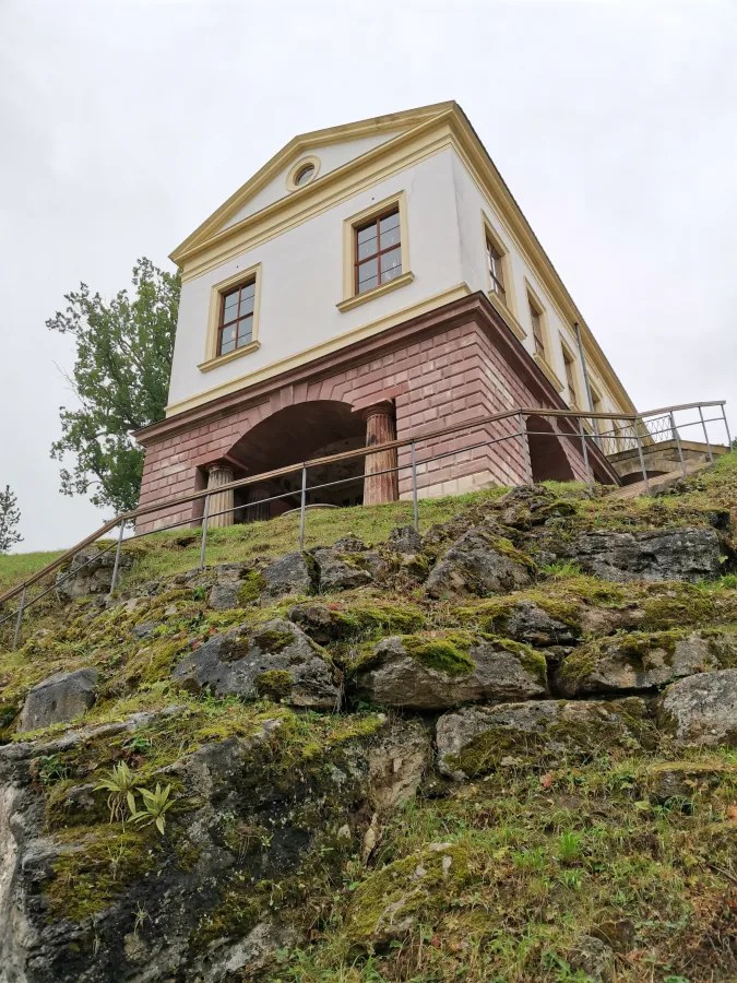 Historisches Gebäude auf einer Anhöhe, umgeben von Bäumen und Felsen.
