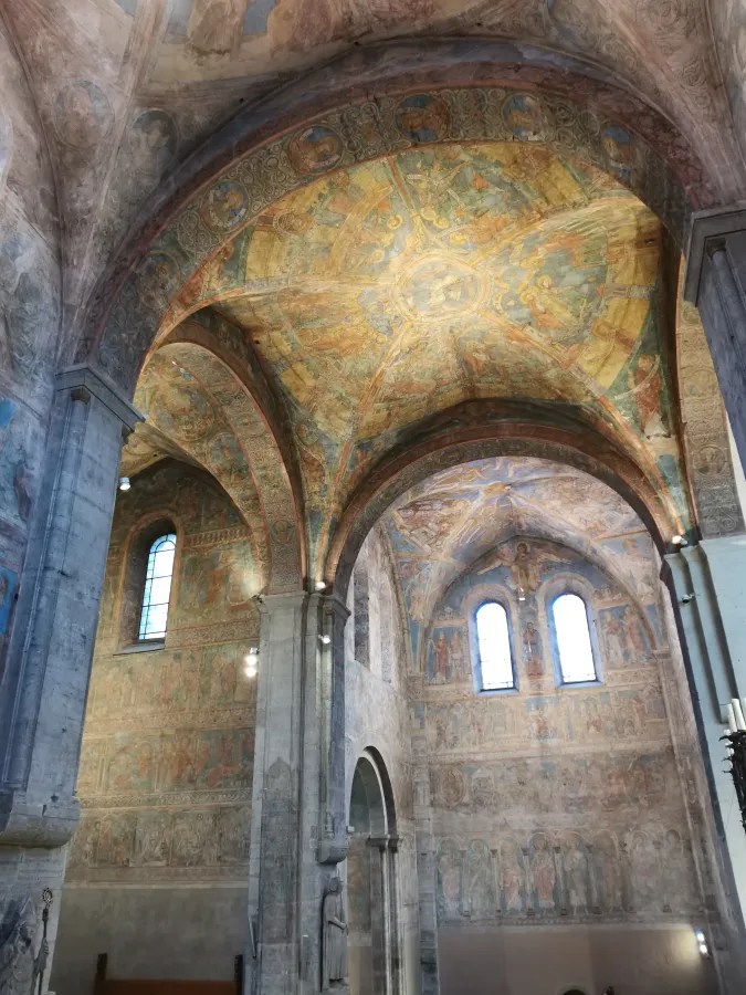 Innenansicht einer historischen Kirche mit buntem, detailliertem Deckengemälde und vielen angestrichenen Wänden.