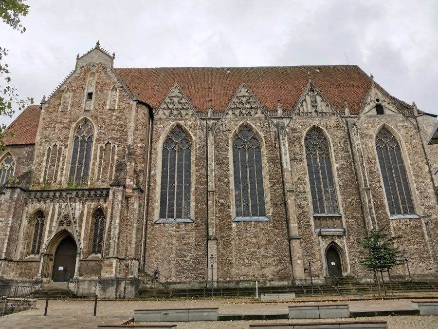Seitenansicht einer historischen Kirche mit gotischen Fenstern und Ziegelbedachung, umgeben von einer ruhigen Landschaft.