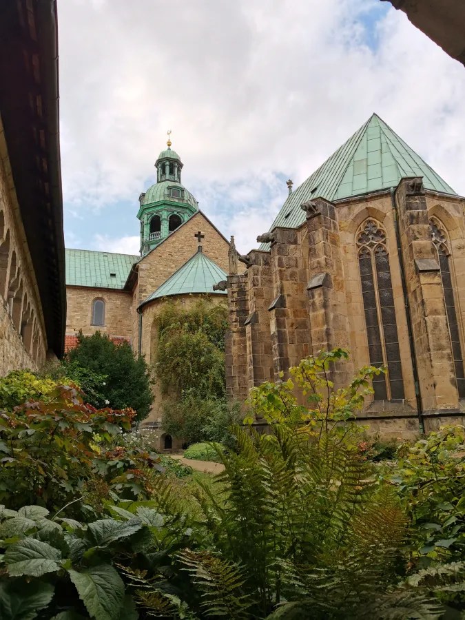 A view of a historic church with a green-topped dome and intricate stone architecture, surrounded by lush greenery and plants.