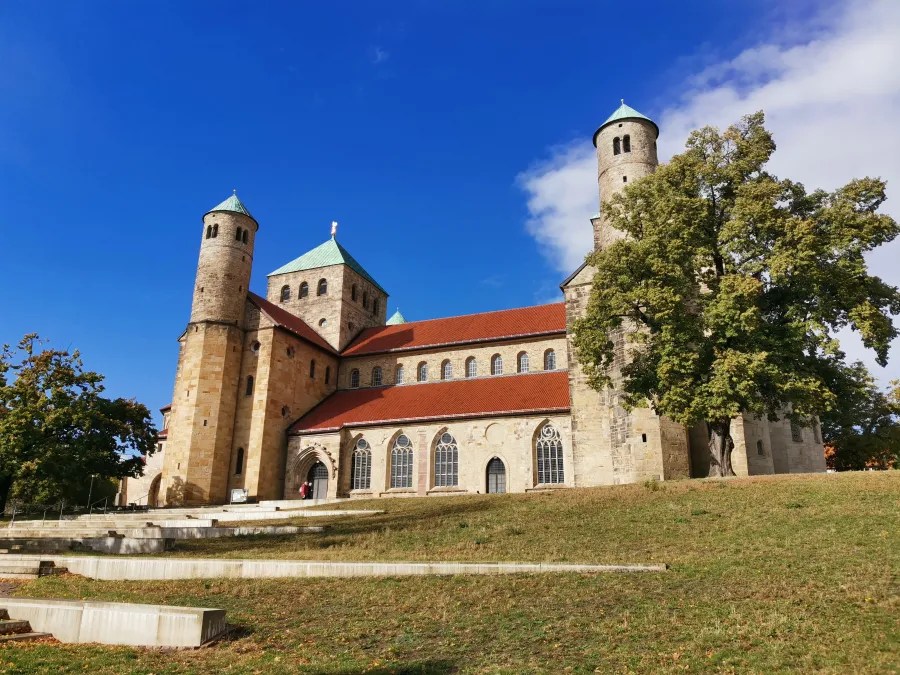 Historisches Kirchengebäude mit zwei Türmen und roten Dächern, umgeben von grünem Gras und Bäumen, vor einem blauen Himmel.
